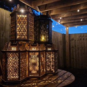 May include: A collection of intricately designed, wooden lanterns with warm interior lighting. The lanterns are stacked on a round wooden table, creating a cozy outdoor ambiance. The background includes a wooden fence and string lights.