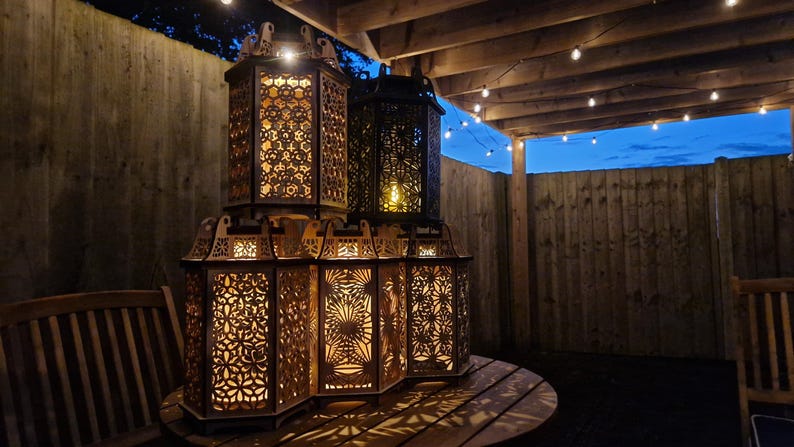May include: A collection of intricately designed, wooden lanterns with warm interior lighting. The lanterns are stacked on a round wooden table, creating a cozy outdoor ambiance. The background includes a wooden fence and string lights.