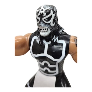 May include: A black and white wrestling action figure with a skull-like mask. The figure is wearing a black and white wrestling suit with a white belt.