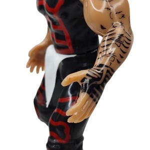 Puede incluir: Figura de acci&oacute;n negra y roja de un luchador enmascarado. La figura tiene tatuajes detallados en el brazo y viste un atuendo negro con detalles rojos. La figura est&aacute; posada en una postura din&aacute;mica, lista para la acci&oacute;n.