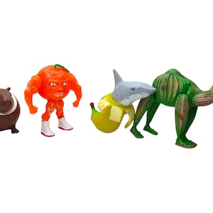 Op de afbeelding: Een verzameling grillige beeldjes, waaronder een bruin nijlpaard, een oranje gespierde figuur, een haai-banaan hybride en een groene kameel. De figuren zijn felgekleurd en hebben overdreven kenmerken, wat een speelse en unieke esthetiek creëert.