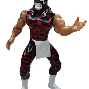 Puede incluir: Figura de acci&oacute;n negra y roja de un luchador enmascarado. La figura tiene una m&aacute;scara negra con detalles blancos, un atuendo negro y rojo y botas blancas. La figura posa con un brazo levantado.