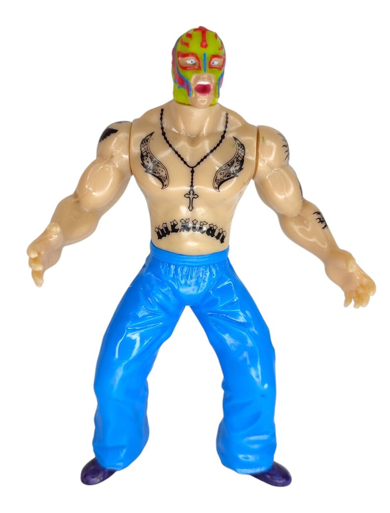 May include: A plastic action figure of a wrestler wearing a yellow, red, and blue mask with a black and white tattoo on his chest. The figure is wearing blue pants and has a black necklace with a cross pendant.