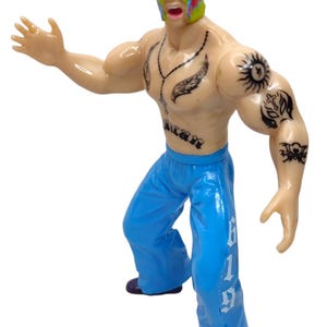 May include: A plastic action figure of a wrestler wearing a colorful mask and blue pants with the number 619 printed on the side. The figure has tattoos on its chest and arms.