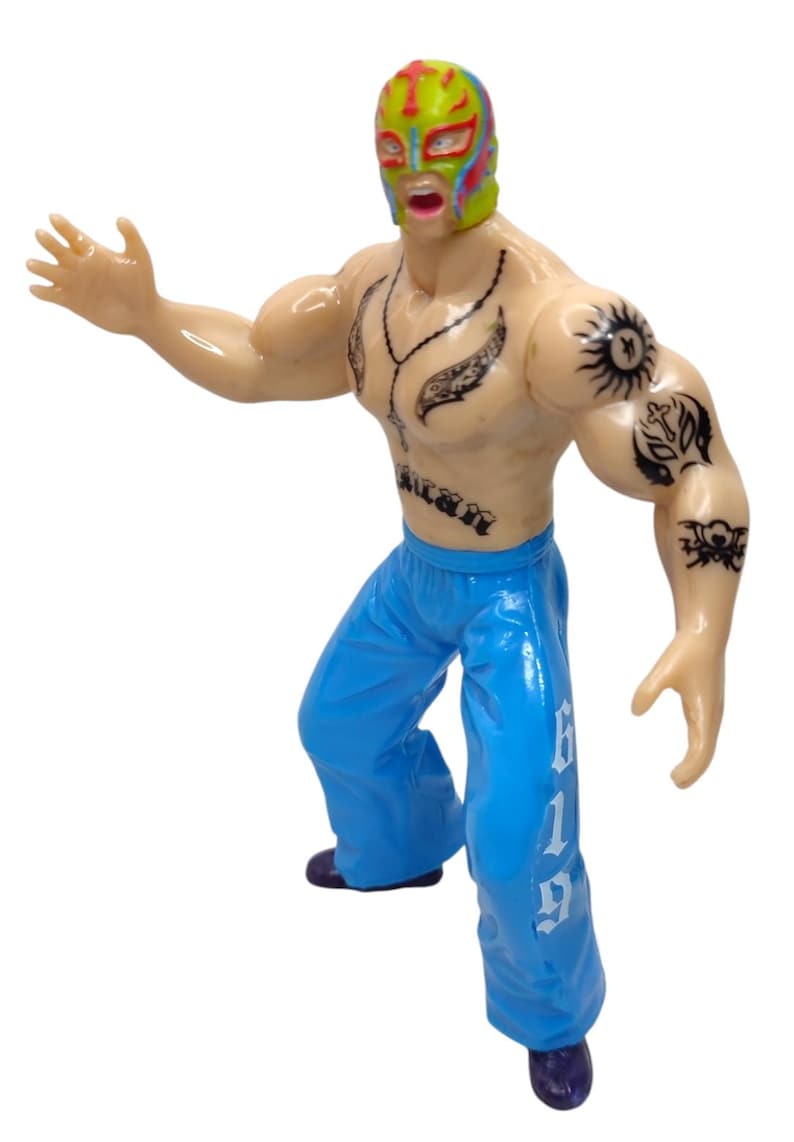 May include: A plastic action figure of a wrestler wearing a colorful mask and blue pants with the number 619 printed on the side. The figure has tattoos on its chest and arms.