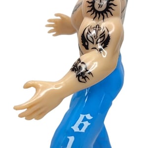 May include: A wrestling action figure with a yellow and red mask, black tattoos, and blue pants with white "619" lettering.