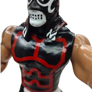 Puede incluir: Figura de acci&oacute;n negra y roja de un luchador enmascarado. La figura tiene una m&aacute;scara negra con detalles blancos y un dise&ntilde;o de calavera. El atuendo del luchador es negro con ribetes rojos. La figura tiene una tela blanca alrededor de la cintura.