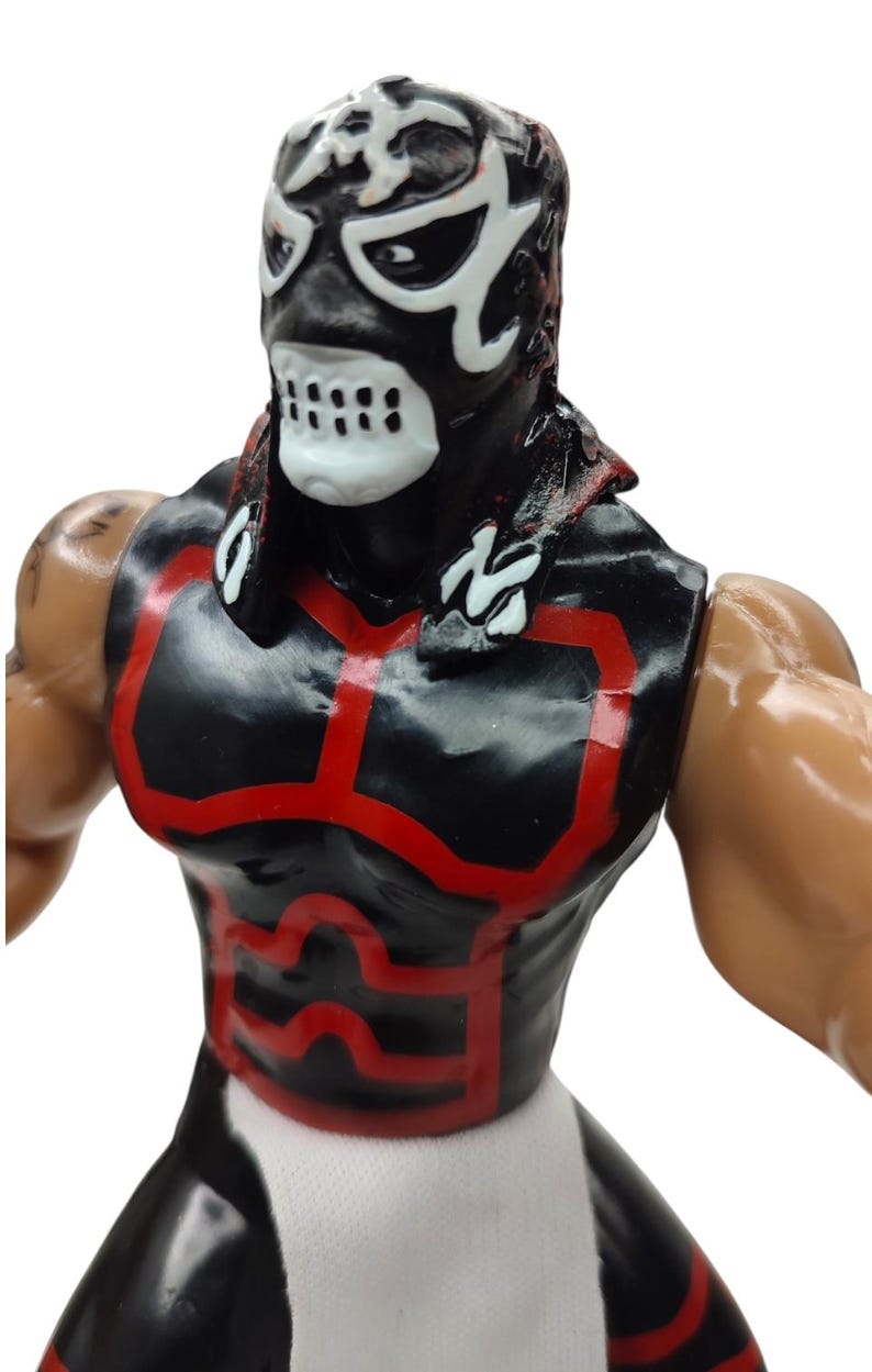 Puede incluir: Figura de acci&oacute;n negra y roja de un luchador enmascarado. La figura tiene una m&aacute;scara negra con detalles blancos y un dise&ntilde;o de calavera. El atuendo del luchador es negro con ribetes rojos. La figura tiene una tela blanca alrededor de la cintura.