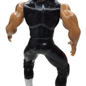 Puede incluir: Una figura de acci&oacute;n de pl&aacute;stico de un personaje musculoso, vista desde atr&aacute;s. La figura viste un atuendo negro con detalles rojos y botas blancas. Tambi&eacute;n es visible una cubierta para la cabeza roja y negra.