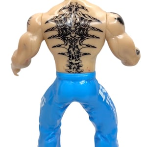 May include: A wrestling action figure with a green mask, black tattoos, and blue pants with white lettering. The figure is posed with its arms outstretched.