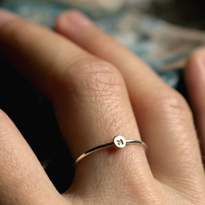 May include: A delicate gold ring with a small round charm featuring the letter "M" on a person's finger.
