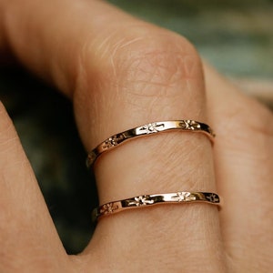 May include: Two thin gold rings with a delicate floral design. The rings are stacked on a finger.