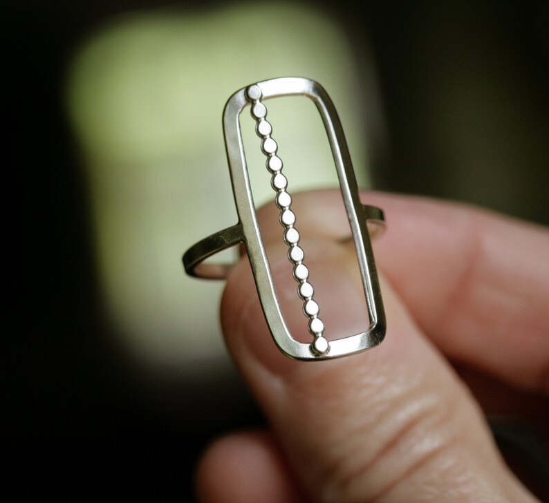 K&ouml;nnte beinhalten: Ein silberner Ring mit einem rechteckigen Design, das eine Reihe kleiner Kreise in der Mitte aufweist.