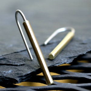 May include: A pair of gold and silver earrings with a simple, minimalist design. The earrings are made of a thin, cylindrical rod with a hook at the top. The rod is made of gold, and the hook is made of silver.