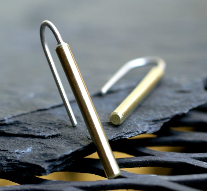 May include: A pair of gold and silver earrings with a simple, minimalist design. The earrings are made of a thin, cylindrical rod with a hook at the top. The rod is made of gold, and the hook is made of silver.