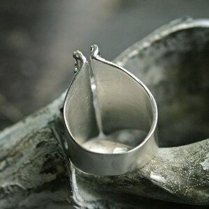 May include: A silver ring with a unique open design. The ring is made of two pieces of metal that are joined together at the top. The ring is sitting on a textured surface.