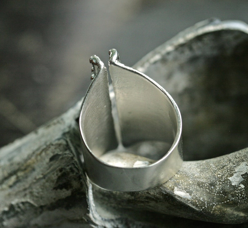 May include: A silver ring with a unique open design. The ring is made of two pieces of metal that are joined together at the top. The ring is sitting on a textured surface.