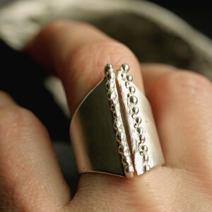 May include: A silver ring with a unique design featuring two vertical rows of beads on either side of a smooth, flat band.