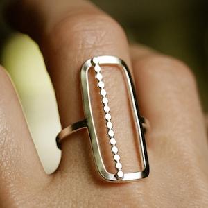 K&ouml;nnte beinhalten: Ein silberner Ring mit einem rechteckigen Design, das eine vertikale Reihe kleiner Kreise aufweist.