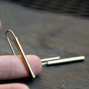 May include: A pair of gold bar earrings with a simple, minimalist design. The earrings are made of a thin, straight bar of gold that is slightly curved at the top to create a hook.