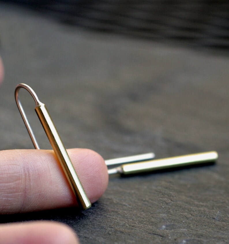 May include: A pair of gold bar earrings with a simple, minimalist design. The earrings are made of a thin, straight bar of gold that is slightly curved at the top to create a hook.