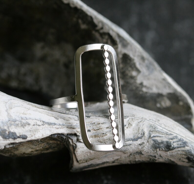 K&ouml;nnte beinhalten: Ein silberner Ring mit einem rechteckigen Design, das eine vertikale Reihe kleiner Kreise aufweist.