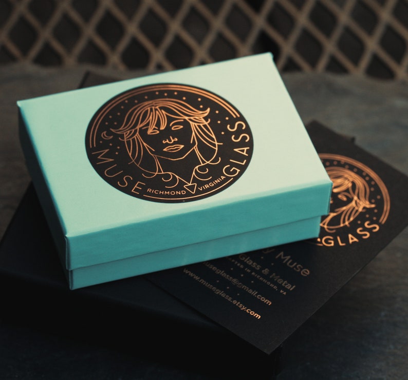 May include: A teal blue box with a gold logo featuring a woman's face and the text "Muse Glass Richmond Virginia". The box is sitting on top of a black box with the text "Muse Glass & Metal Richmond, VA www.museglass.etsy.com museglass@gmail.com" in gold lettering.