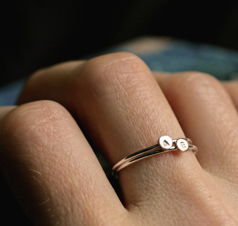 May include: A delicate gold ring with two small round charms, one with the letter "A" and the other with the letter "B", worn on a finger.