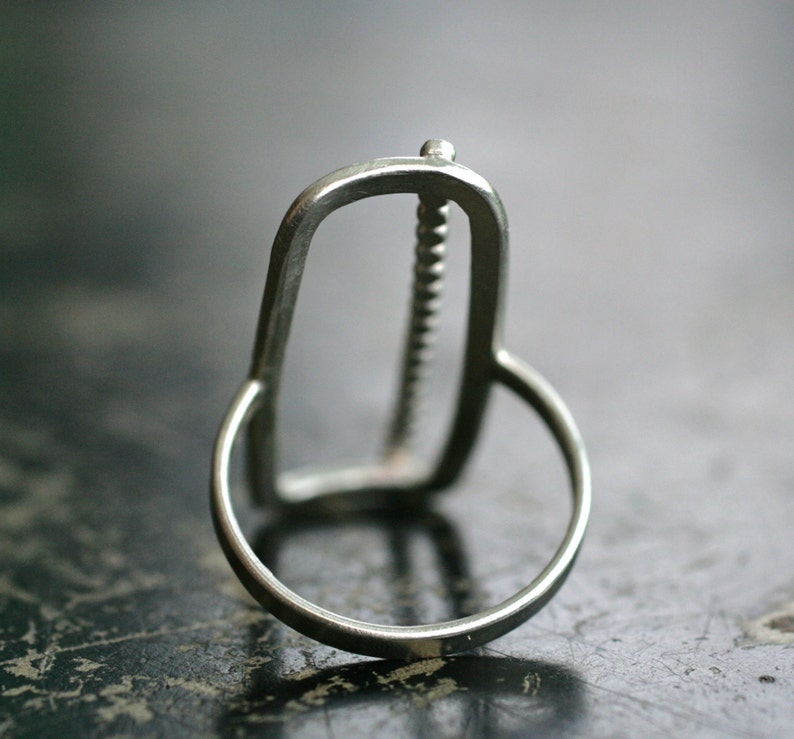K&ouml;nnte beinhalten: Ein silberner Ring mit einem einzigartigen, abstrakten Design. Der Ring verf&uuml;gt &uuml;ber eine strukturierte, vertikale Leiste in einem runden Rahmen.