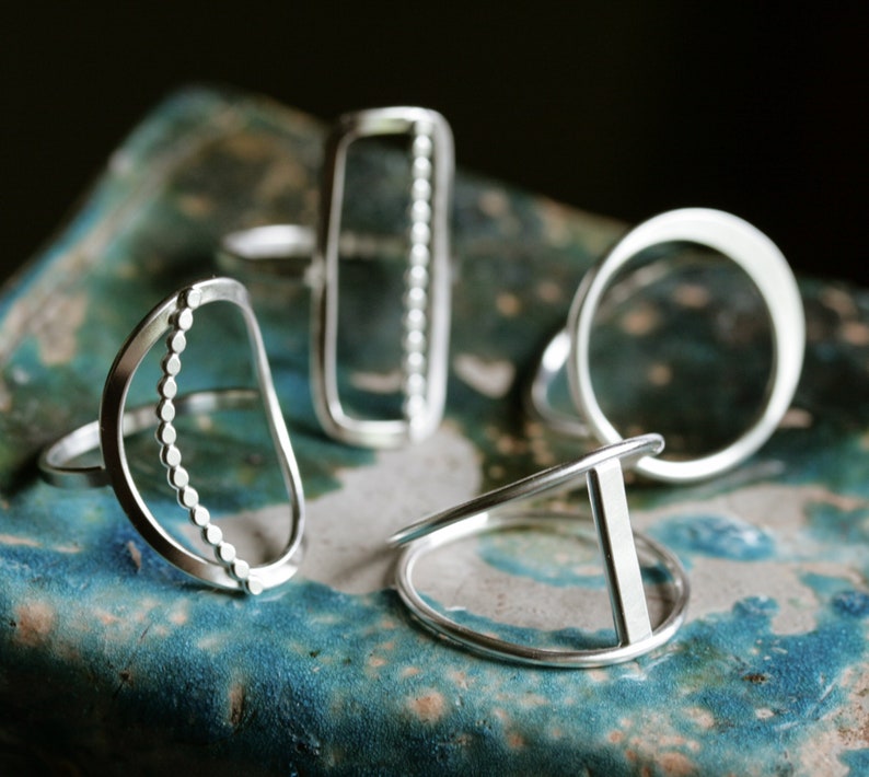 K&ouml;nnte beinhalten: Vier silberne Ringe mit verschiedenen geometrischen Formen. Die Ringe liegen auf einer blauen und gr&uuml;nen gekachelten Oberfl&auml;che.