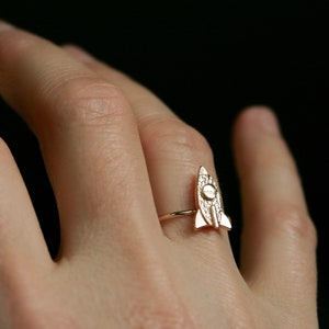 May include: A gold-toned ring with a rocket ship design. The ring is worn on a person's finger.