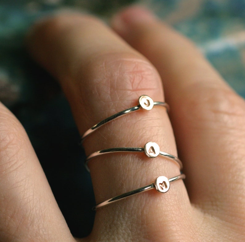 May include: Three delicate gold rings with initial charms. The rings are stacked on a finger, with the letters J, A, and M visible.