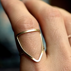 May include: A gold-toned geometric ring with a triangular shape. The ring is worn on a finger.