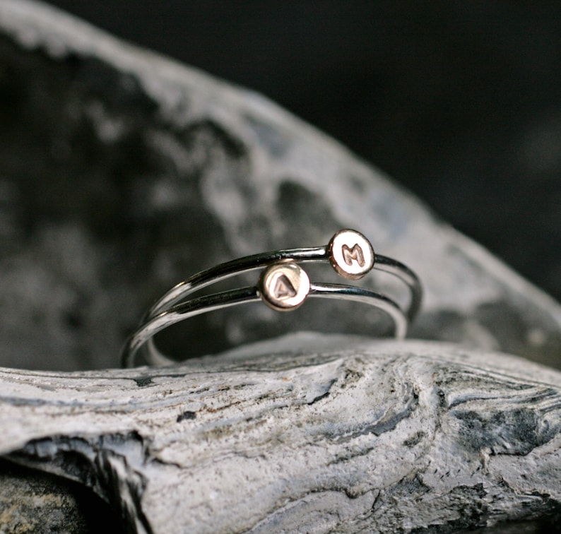 May include: Two silver rings with gold accents. The rings are stacked on top of each other. The top ring has the letter "M" engraved on it, and the bottom ring has the letter "A" engraved on it.