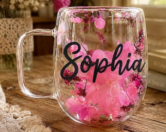 Könnte beinhalten: Ein transparenter doppelwandiger Glasbecher mit Henkel und dem in schwarzer Schreibschrift aufgedruckten Namen Sophia. Der Zwischenraum der Glaswände ist mit leuchtend rosa getrockneten Blütenblättern gefüllt, die ein florales Design bilden.