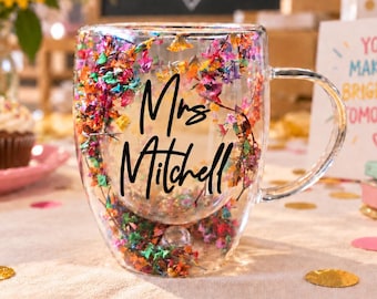 Könnte beinhalten: Ein transparenter doppelwandiger Glasbecher mit Henkel und einer dekorativen Schicht aus bunten getrockneten Blütenblättern zwischen den Wänden. Der Text "Mrs Mitchell" ist in schwarzer Schreibschrift auf der Vorderseite aufgedruckt.