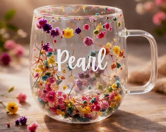 Könnte beinhalten: Ein klarer Glasbecher mit Henkel, in dessen Glaswände kleine, bunte Trockenblumen eingearbeitet sind. Das Wort Pearl ist in weißer Schreibschrift mittig auf der Vorderseite des Bechers aufgedruckt, umgeben von rosa, gelben und blauen Blüten.