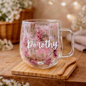 K&ouml;nnte beinhalten: Ein transparenter doppelwandiger Glasbecher mit eingebetteten rosa und violetten Trockenblumen zwischen den Schichten. Der Name Dorothy ist in wei&szlig;er Schreibschrift auf der Vorderseite des Bechers aufgedruckt. Ein kleiner Holzl&ouml;ffel liegt auf einem quadratischen Bambusuntersetzer.