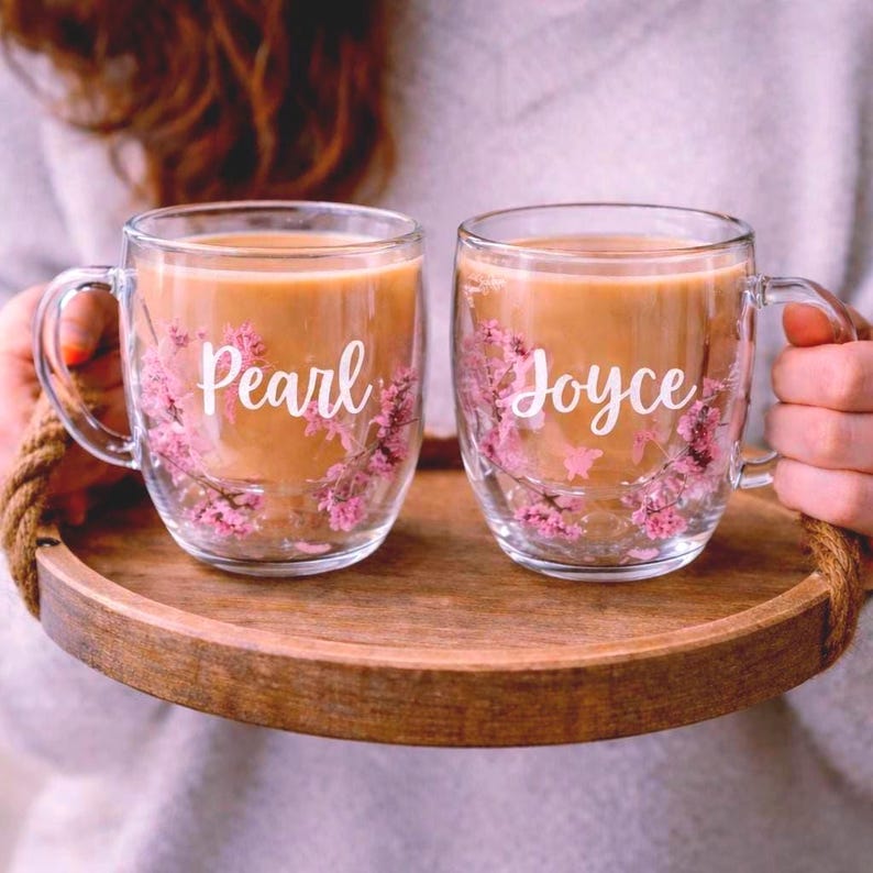K&ouml;nnte beinhalten: Zwei durchsichtige Glastassen gef&uuml;llt mit einem beigen Getr&auml;nk, verziert mit eingebetteten rosa Bl&uuml;ten und wei&szlig;er Schreibschrift. Die linke Tasse tr&auml;gt den Namen Pearl, die rechte den Namen Joyce, beide stehen auf einem runden Holztablett.