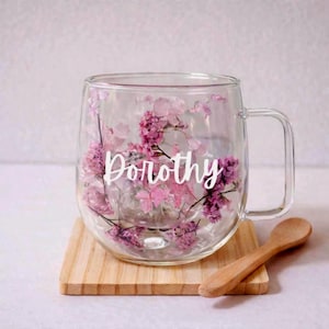 K&ouml;nnte beinhalten: Ein klarer Glasbecher mit doppelwandigem Design, gef&uuml;llt mit gepressten rosa und violetten Blumen. Der Name Dorothy ist in wei&szlig;er Schreibschrift in der Mitte des Glases aufgedruckt. Der Becher hat einen stabilen Henkel und eine runde Form.
