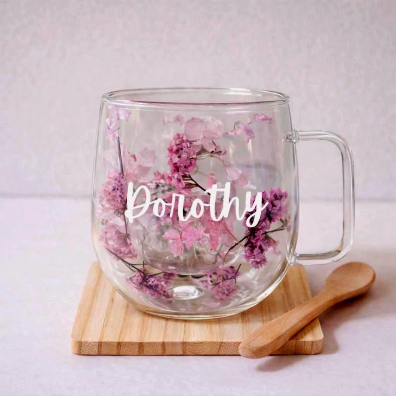 K&ouml;nnte beinhalten: Ein klarer Glasbecher mit doppelwandigem Design, gef&uuml;llt mit gepressten rosa und violetten Blumen. Der Name Dorothy ist in wei&szlig;er Schreibschrift in der Mitte des Glases aufgedruckt. Der Becher hat einen stabilen Henkel und eine runde Form.