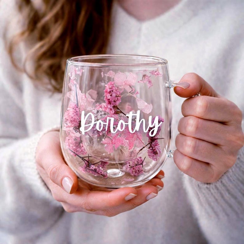 K&ouml;nnte beinhalten: Ein doppelwandiger Becher aus klarem Glas, der zwischen den Schichten gepresste rosa und violette Blumen enth&auml;lt. Der Name Dorothy ist in wei&szlig;er Schreibschrift auf der Vorderseite des Glases aufgedruckt und mittig zwischen den Blumen platziert.