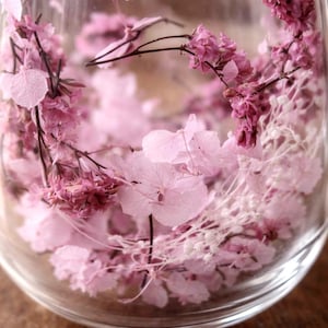 K&ouml;nnte beinhalten: Ein durchsichtiges Glasgef&auml;&szlig; gef&uuml;llt mit einer Auswahl an konservierten Blumen in verschiedenen Rosa- und Wei&szlig;t&ouml;nen. Das Arrangement besteht aus zarten Hortensienbl&uuml;tenbl&auml;ttern, kleinen Zweigen getrockneter Bl&uuml;ten und d&uuml;nnen Stielen.