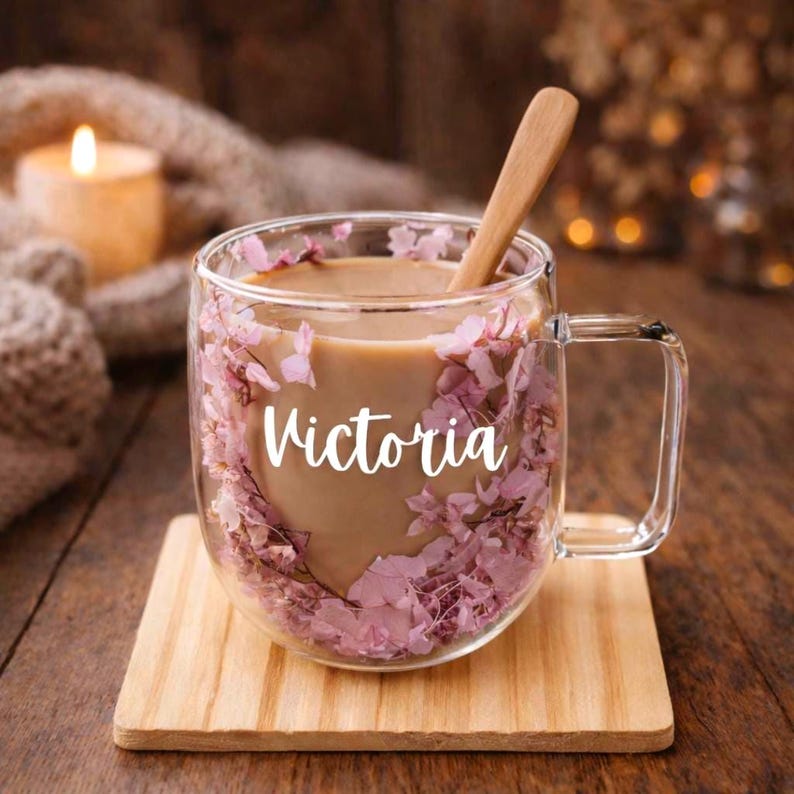 K&ouml;nnte beinhalten: Ein doppelwandiger Glasbecher gef&uuml;llt mit einem hellbraunen Getr&auml;nk und zwischen den W&auml;nden eingebetteten rosa Bl&uuml;tenbl&auml;ttern. Der Name Victoria ist in wei&szlig;er Schreibschrift auf der Vorderseite aufgedruckt. Ein Holzl&ouml;ffel steckt im Becher.