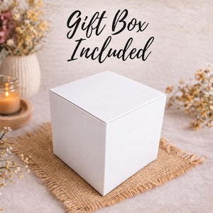 K&ouml;nnte beinhalten: Eine schlichte, quadratische, wei&szlig;e Geschenkbox mit glatter, matter Oberfl&auml;che. Der Text Gift Box Included ist in schwarzer Schreibschrift &uuml;ber der Box aufgedruckt. Die Box steht auf einem quadratischen St&uuml;ck braunem Jute-Stoff mit ausgefransten R&auml;ndern.