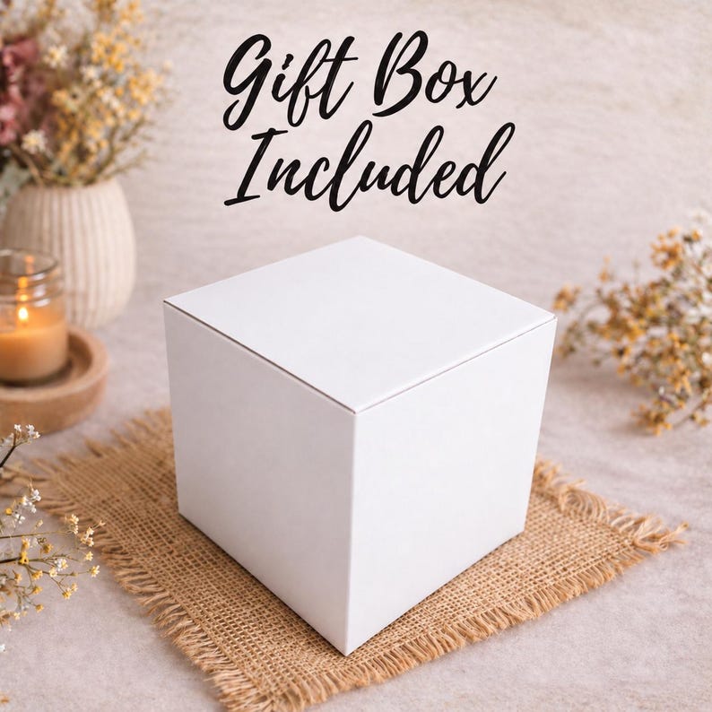 K&ouml;nnte beinhalten: Eine schlichte, quadratische, wei&szlig;e Geschenkbox mit glatter, matter Oberfl&auml;che. Der Text Gift Box Included ist in schwarzer Schreibschrift &uuml;ber der Box aufgedruckt. Die Box steht auf einem quadratischen St&uuml;ck braunem Jute-Stoff mit ausgefransten R&auml;ndern.
