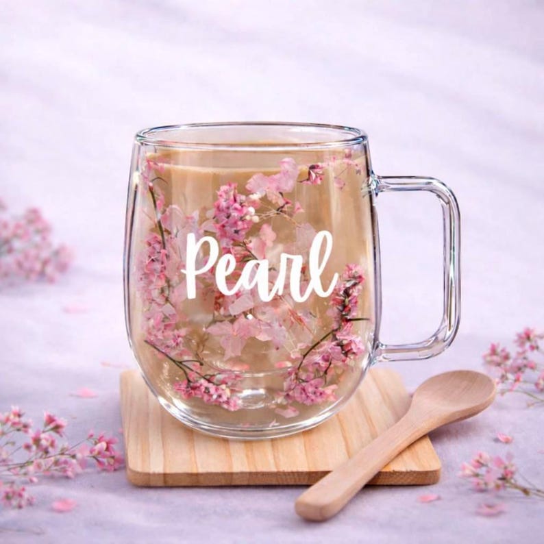 K&ouml;nnte beinhalten: Ein klarer Glasbecher mit Henkel, gef&uuml;llt mit einem hellbraunen Getr&auml;nk. Das Glas ist mit eingebetteten rosa Trockenblumen und dem in wei&szlig;er Schreibschrift gedruckten Text Pearl auf der Vorderseite versehen. Der Becher steht auf einem Holzuntersetzer.