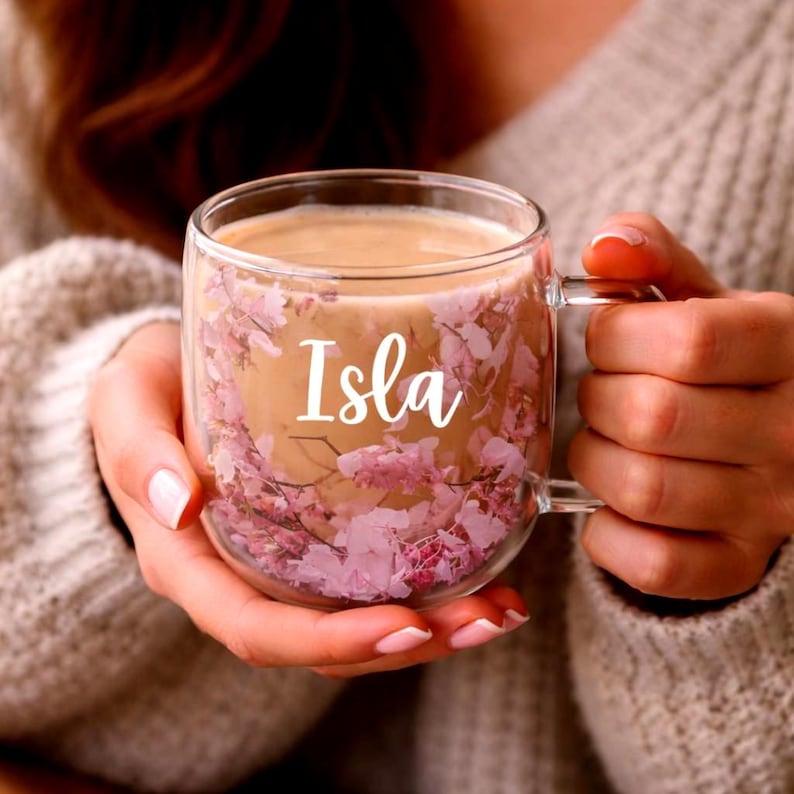 K&ouml;nnte beinhalten: Ein transparenter doppelwandiger Glasbecher mit einer dekorativen Schicht aus kleinen rosa Bl&uuml;tenbl&auml;ttern, die zwischen den Glasw&auml;nden eingebettet sind. Der Name Isla ist in wei&szlig;er Schreibschrift auf der Vorderseite des mit Kaffee gef&uuml;llten Bechers.