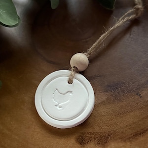 May include: A white, circular ceramic ornament with a stamped chicken design, suspended by a natural twine cord and a wooden bead. The ornament is set against a blurred wooden background, with green foliage in the upper left corner.