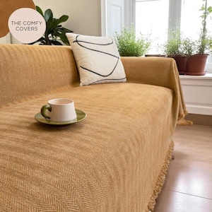 May include: A mustard-colored sofa cover with a herringbone pattern, fringed edges, and a white pillow with black abstract lines. A white mug with a green handle sits on a green saucer. The text "THE COMFY COVERS" is in a circle in the upper left.